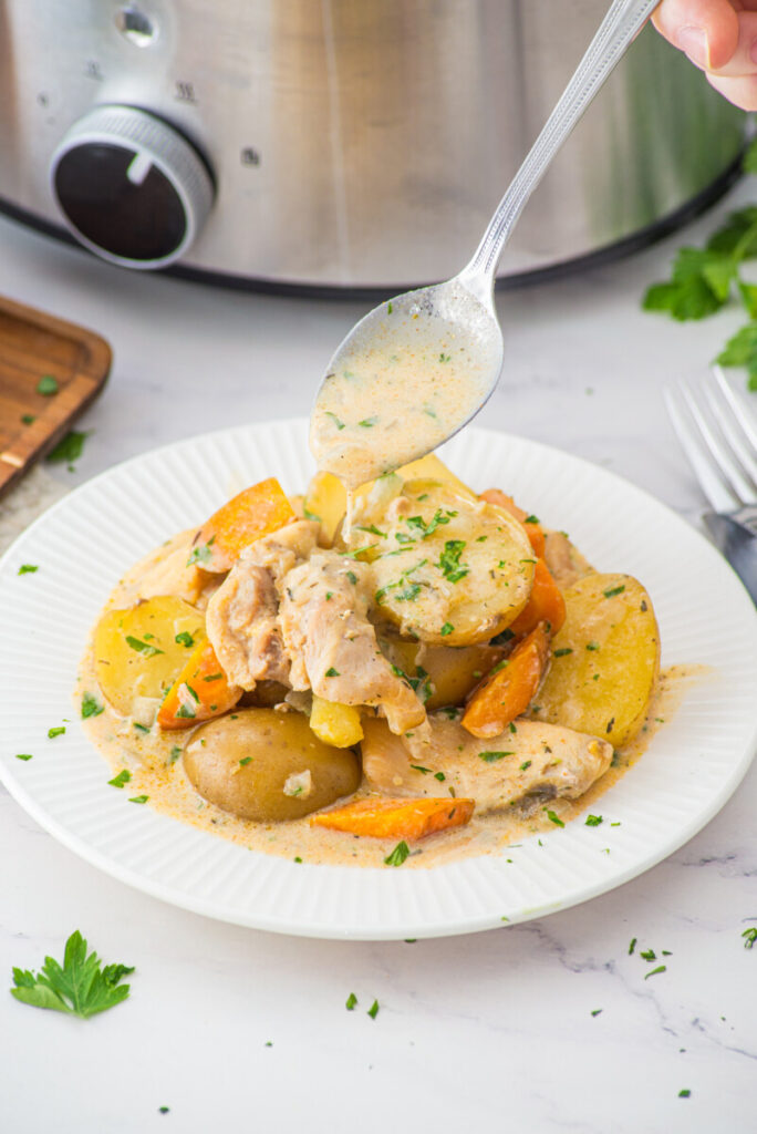 Creamy slow cooker chicken and potatoes with carrots garnished with fresh parsley, plated on white dish with sauce being drizzled