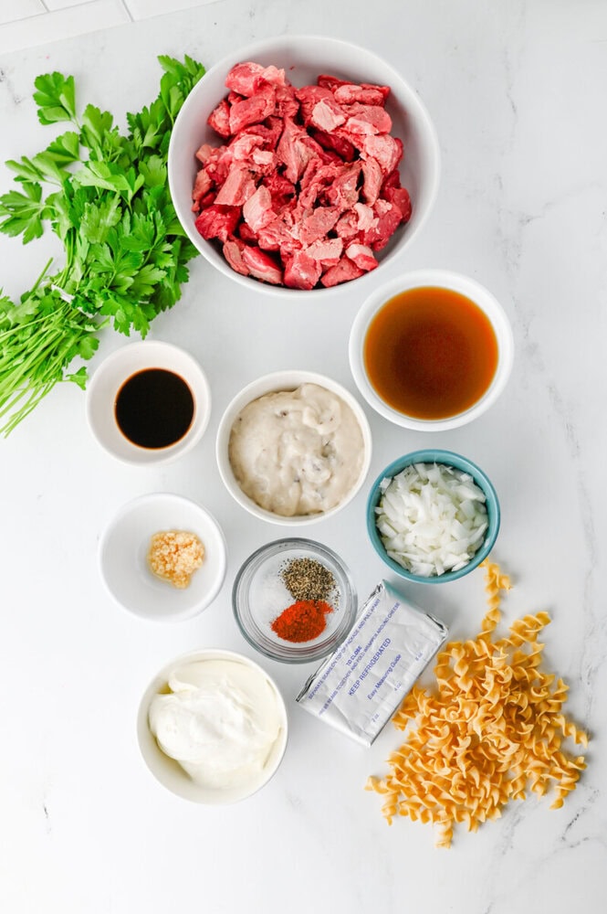 Slow cooker creamy beef and noodles ingredients laid out overhead on marble including raw beef cubes, pasta, sour cream, broth, and fresh herbs