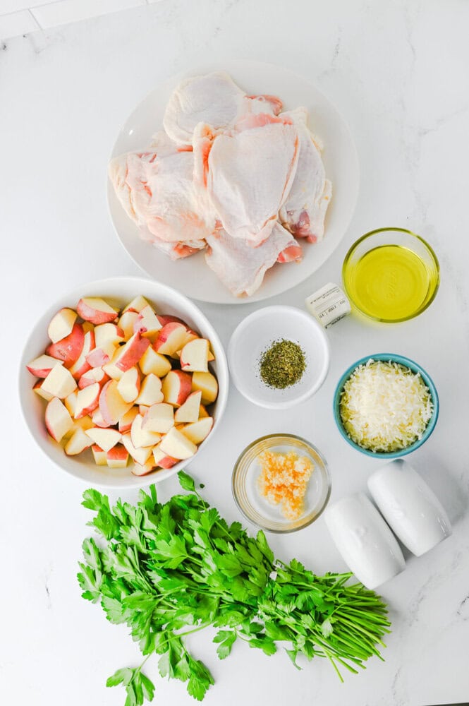 Raw chicken breasts, diced potatoes, fresh parsley, minced garlic, shredded parmesan, olive oil, and seasonings arranged on white plates and bowls for slow cooker recipe preparation