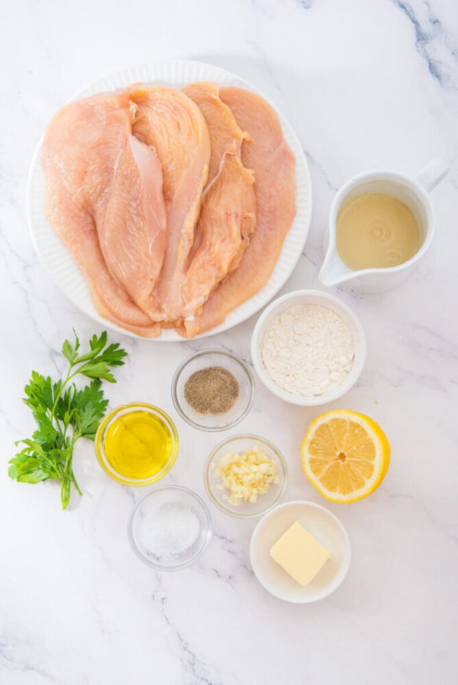 Lemon garlic chicken ingredients laid out flat - raw chicken breasts, lemon, garlic, olive oil, butter, flour, and parsley on marble