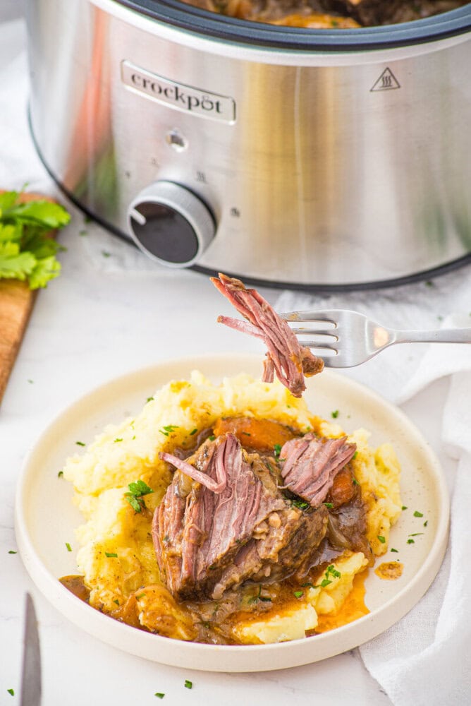 Slow cooker braised short ribs plated over mashed potatoes with Crock-Pot in background