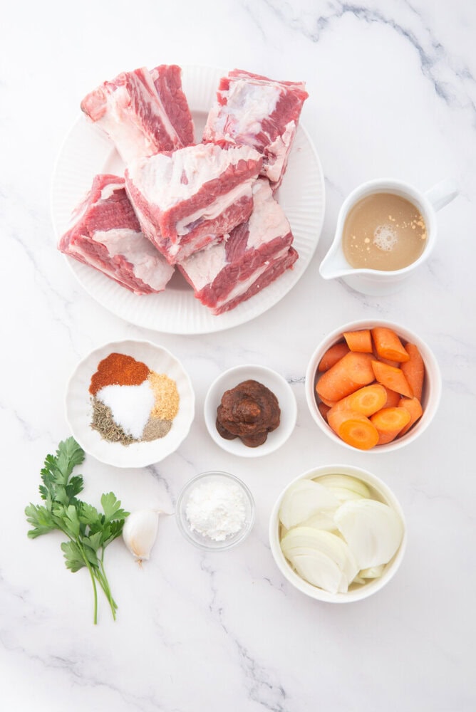 Slow cooker braised short ribs ingredients laid out including raw beef short ribs, carrots, onions, garlic, parsley, spices, and beef broth on marble background