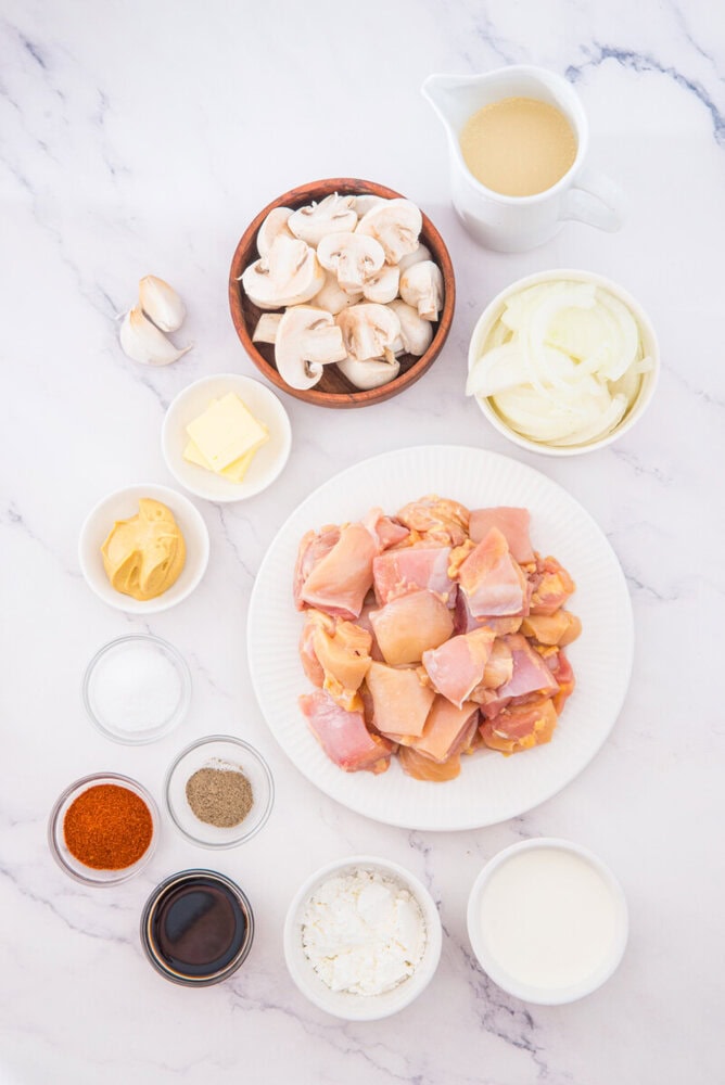 Slow cooker chicken stroganoff ingredients laid out including raw chicken pieces, sliced mushrooms, butter, sour cream, and seasonings on marble countertop