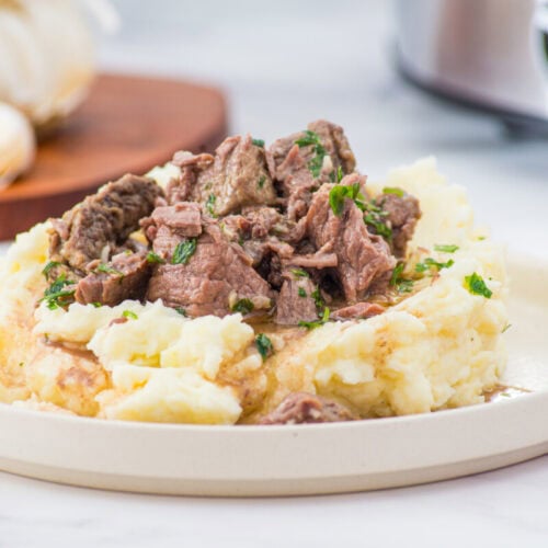 Slow cooker garlic butter steak bites served over mashed potatoes with fresh parsley garnish