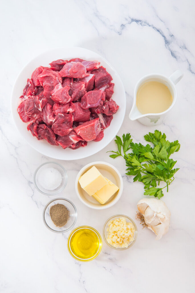 Slow cooker garlic butter steak bites ingredients including cubed beef, butter, fresh parsley, garlic, and seasonings laid out on marble