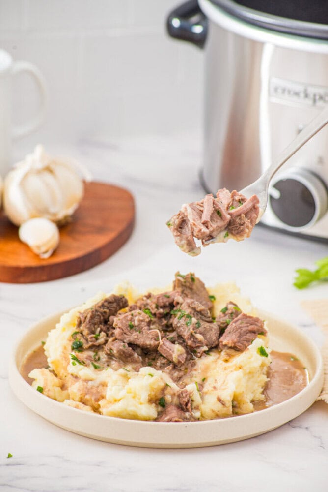 Slow cooker garlic butter steak bites served over mashed potatoes with parsley, fork lifting a bite