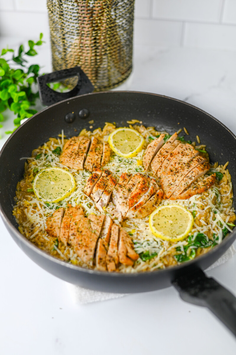 Creamy lemon parmesan chicken orzo in a black skillet with sliced chicken, lemon garnish, and fresh herbs