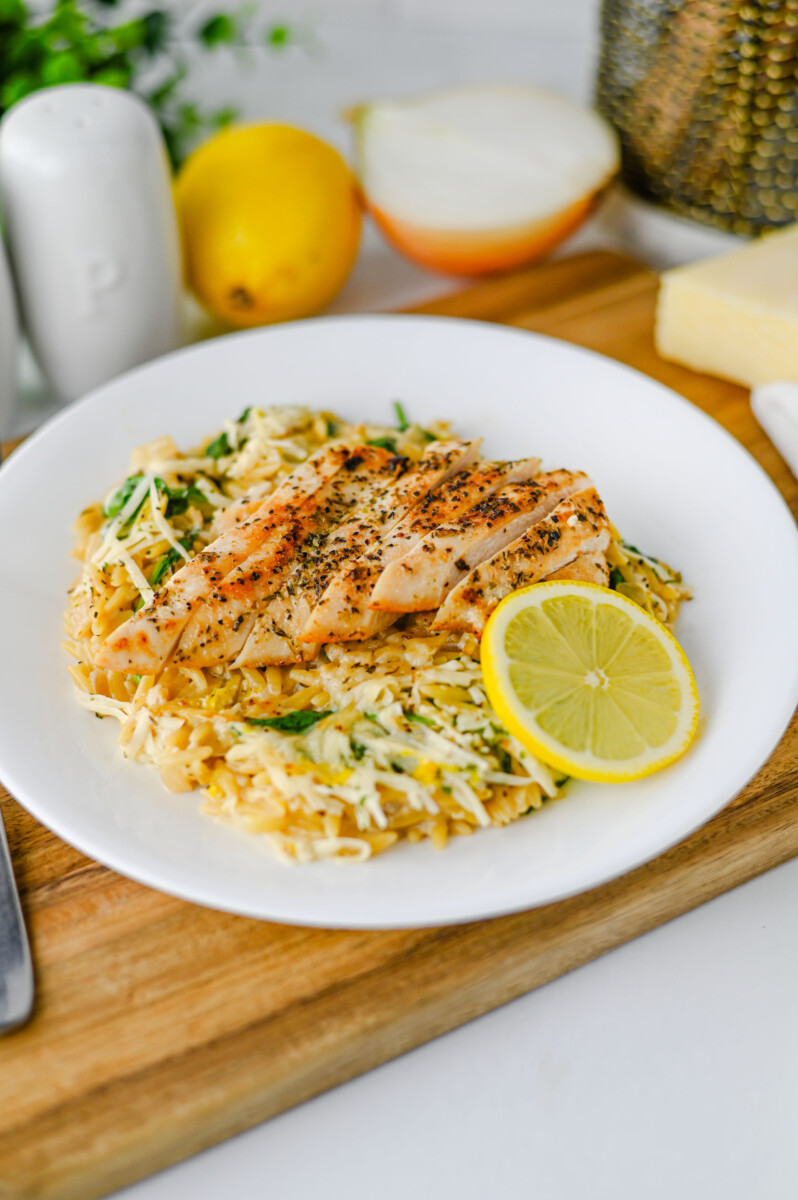 Creamy lemon parmesan chicken orzo with sliced chicken, pasta, and fresh lemon slice on white plate