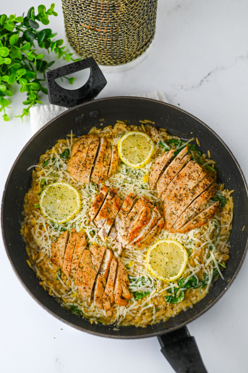 Creamy lemon parmesan chicken orzo in a black skillet with lemon slices and fresh herbs garnish