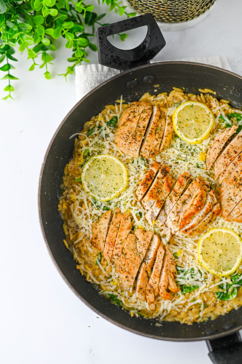 Creamy Lemon Parmesan Chicken Orzo in a black skillet with lemon slices and parmesan cheese garnish