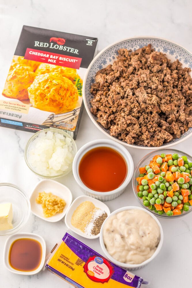 Cheddar Bay Beef Cobbler ingredients including ground beef, Red Lobster biscuit mix, vegetables, and seasonings arranged overhead on marble