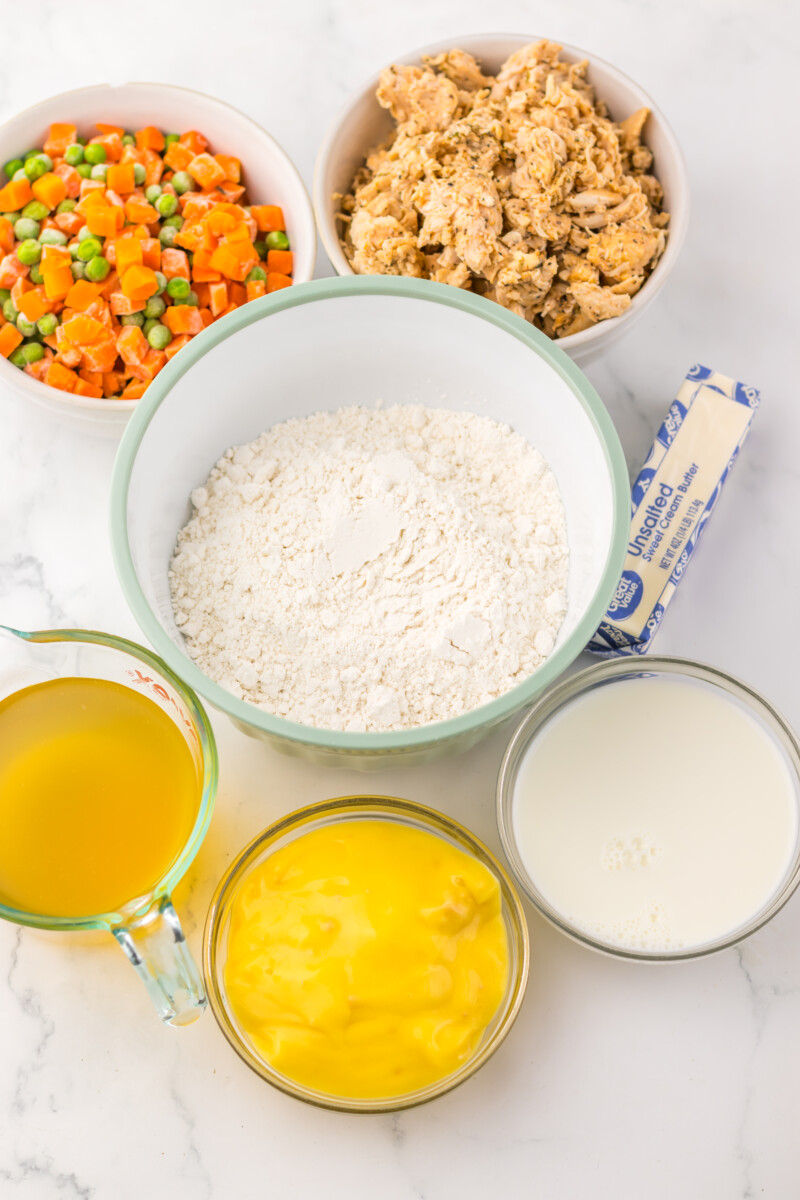 Chicken cobbler ingredients arranged in bowls from above including flour, vegetables, eggs, butter, and milk