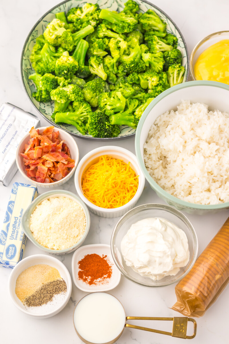 Broccoli rice casserole ingredients laid out including fresh broccoli florets, cooked rice, shredded cheese, bacon bits, sour cream, unsalted butter, and seasonings