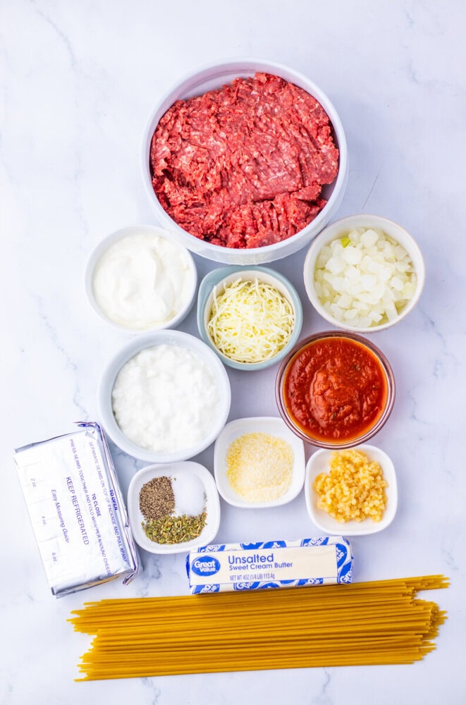 Million Dollar Spaghetti Casserole ingredients laid out in bowls including ground beef, sour cream, cheese, vegetables, and pasta on marble background