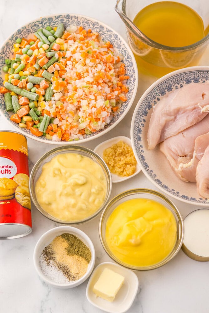 Slow cooker chicken and dumplings pot pie ingredients laid out overhead including diced vegetables, raw chicken, mayo, mustard, broth and seasonings