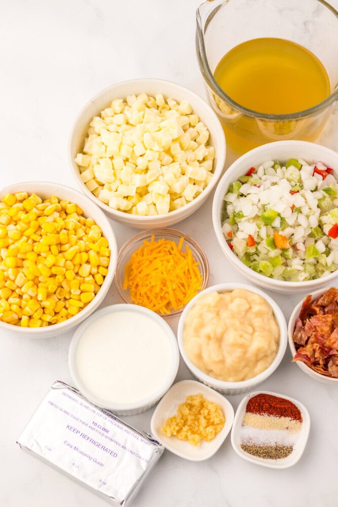 Ingredients for slow cooker creamy bacon corn chowder laid out in bowls and containers including corn, potatoes, vegetables, cheese, mayo, and broth