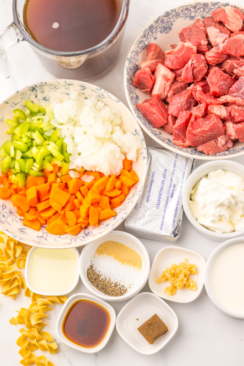 Ingredients for slow cooker creamy beef noodle soup laid out and organized including raw beef, diced onions, peppers, pasta, broth, and seasonings