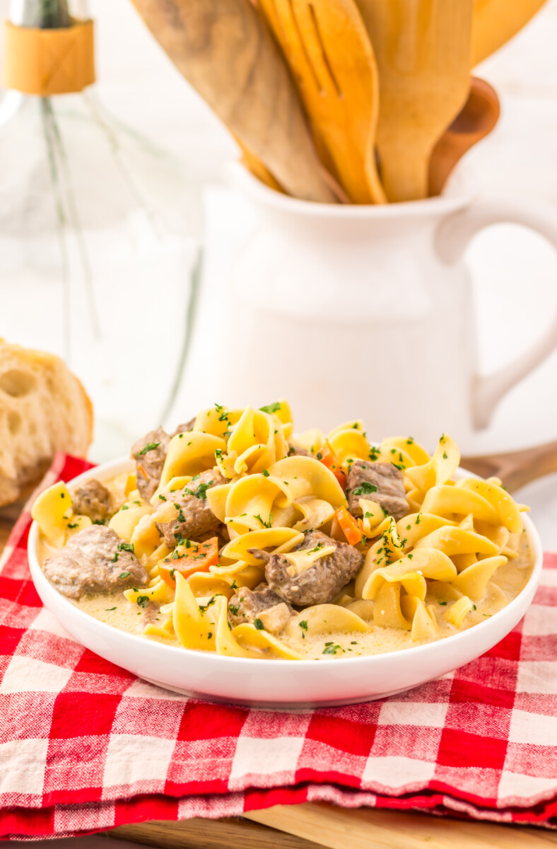 Slow cooker creamy beef noodle soup plated in white bowl with fresh herbs and breadsticks in background