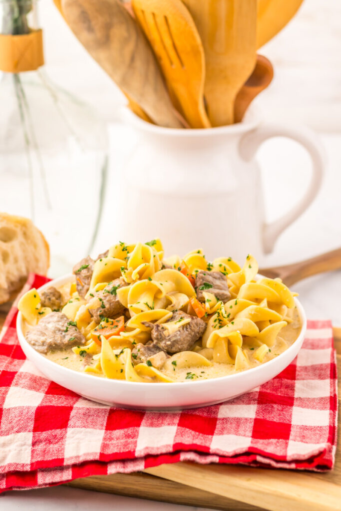 Slow cooker creamy beef noodle soup with farfalle pasta and tender beef in white bowl on checkered napkin