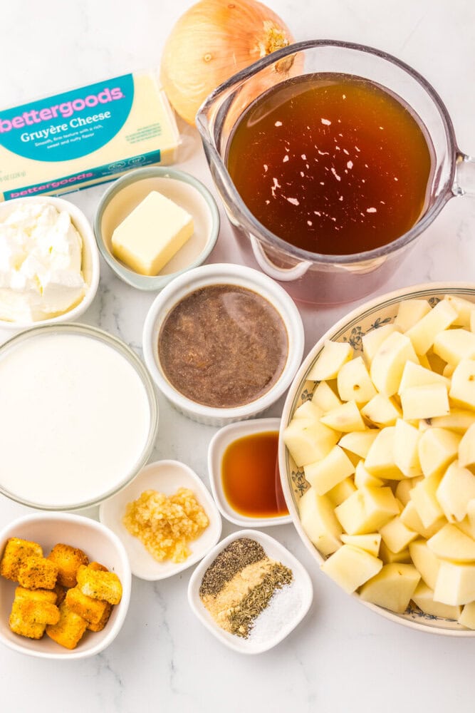 Prepped ingredients for slow cooker French onion potato soup including potatoes, onion, Gruyère cheese, broth, cream, and seasonings arranged in bowls