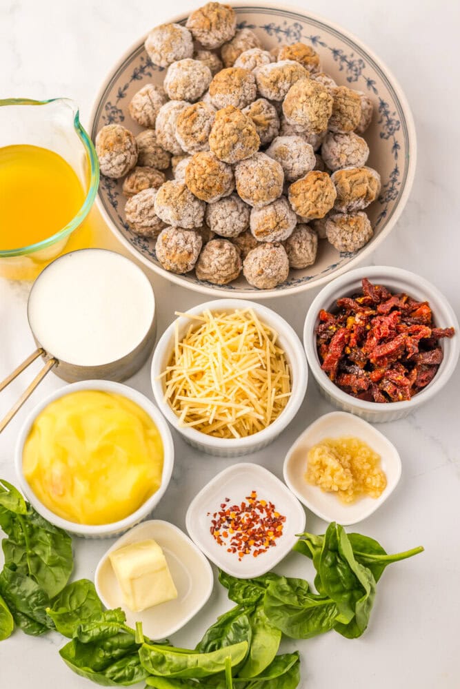 Slow cooker meatball ingredients laid out including cooked meatballs, cream sauce, shredded cheese, bacon bits, garlic, and fresh spinach