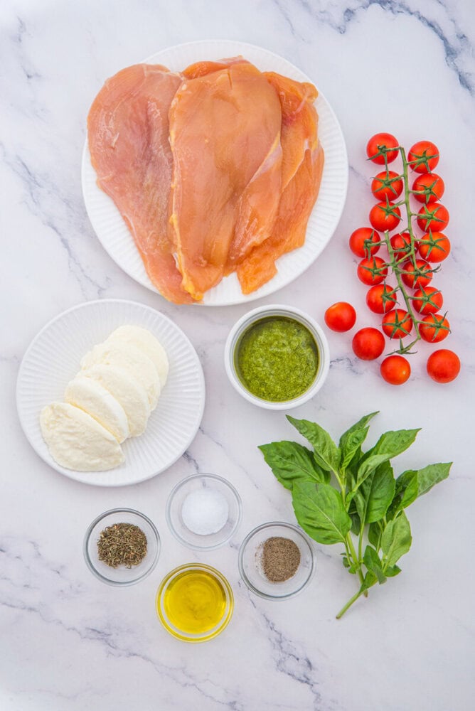 Chicken Margherita ingredients arranged overhead including prosciutto, mozzarella, tomatoes, basil, and pesto on marble surface