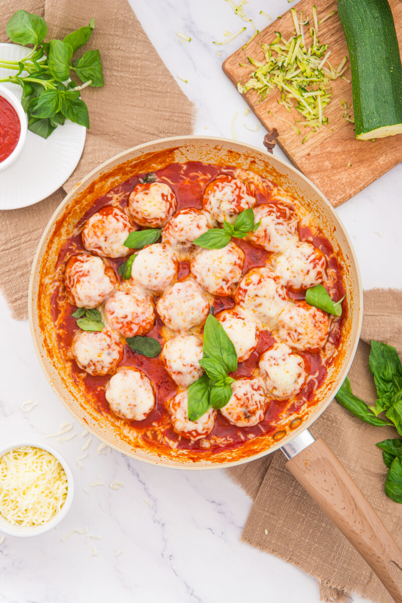 Chicken parmesan meatballs in tomato sauce with melted cheese and basil in a skillet, overhead view with prep ingredients