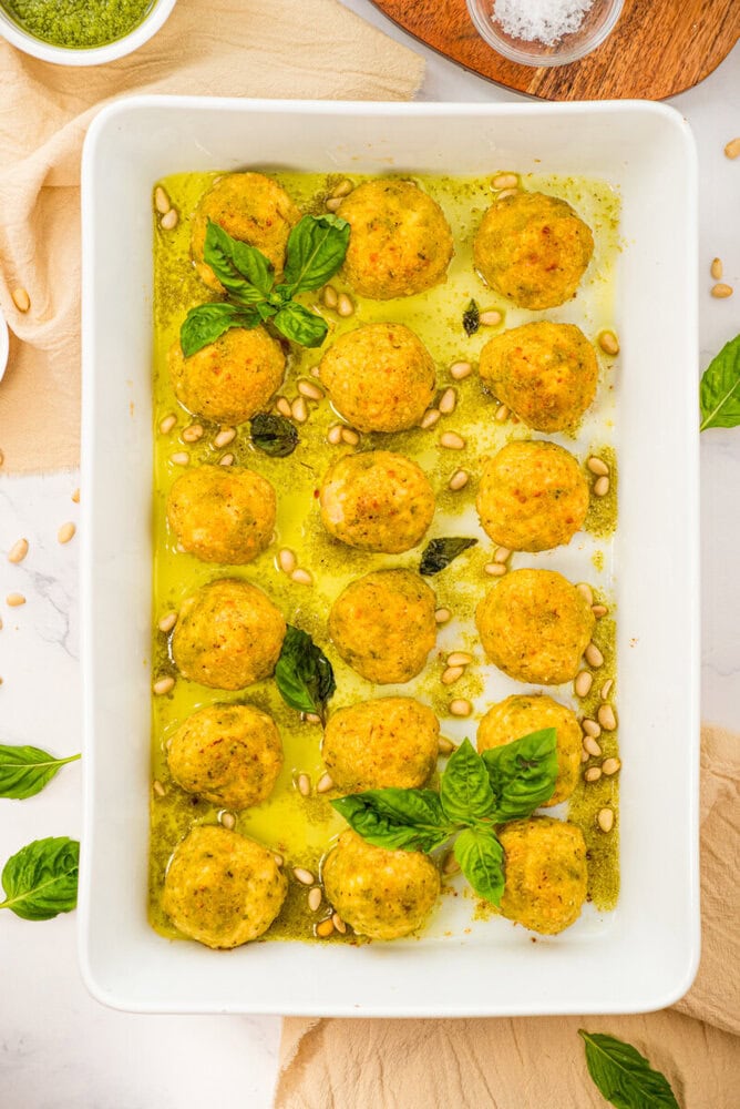 Chicken pesto meatballs in pesto sauce with pine nuts and fresh basil in white baking dish