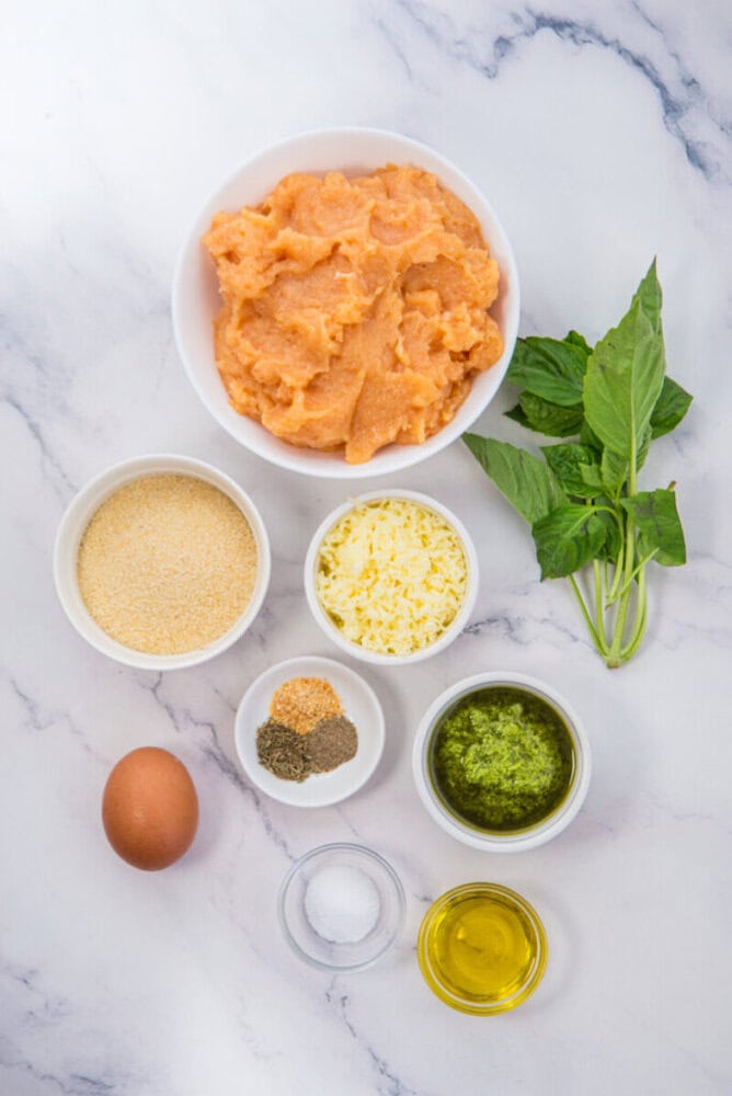 Chicken pesto meatball ingredients arranged in white bowls on marble countertop with fresh basil garnish