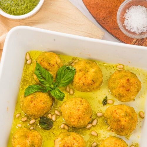 Chicken pesto meatballs in green sauce garnished with basil and pine nuts in a white dish