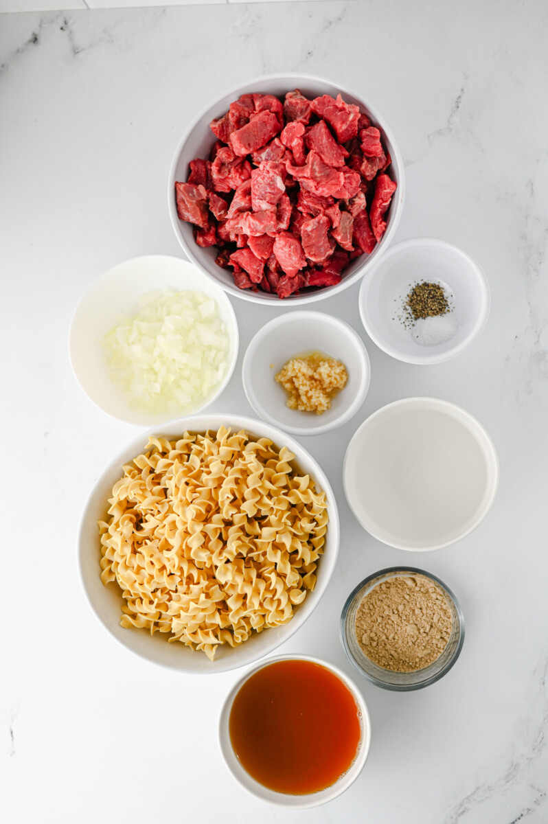 Crockpot Beef and Noodles ingredients laid out in bowls including raw beef cubes, egg noodle pasta, onions, garlic, broth and seasonings