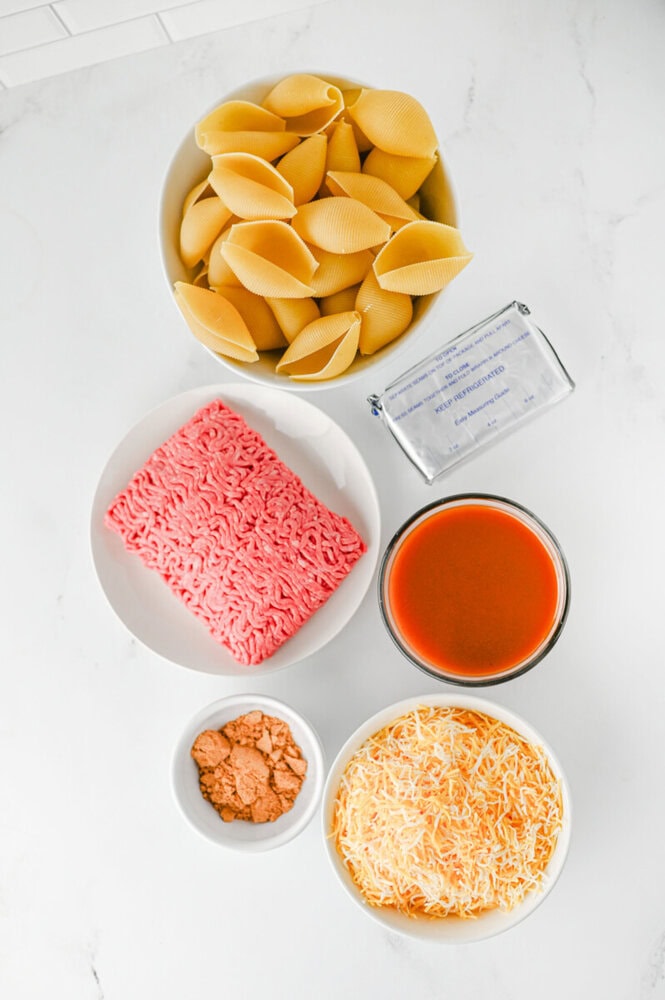 Ingredients for cheesy taco stuffed shells laid out flat including pasta shells, ground beef, tomato sauce, cheese, and seasonings