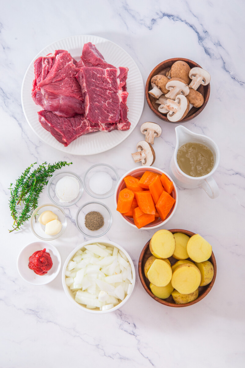Prepped ingredients for slow cooker beef pot roast including raw beef, potatoes, carrots, mushrooms, and seasonings arranged on marble surface