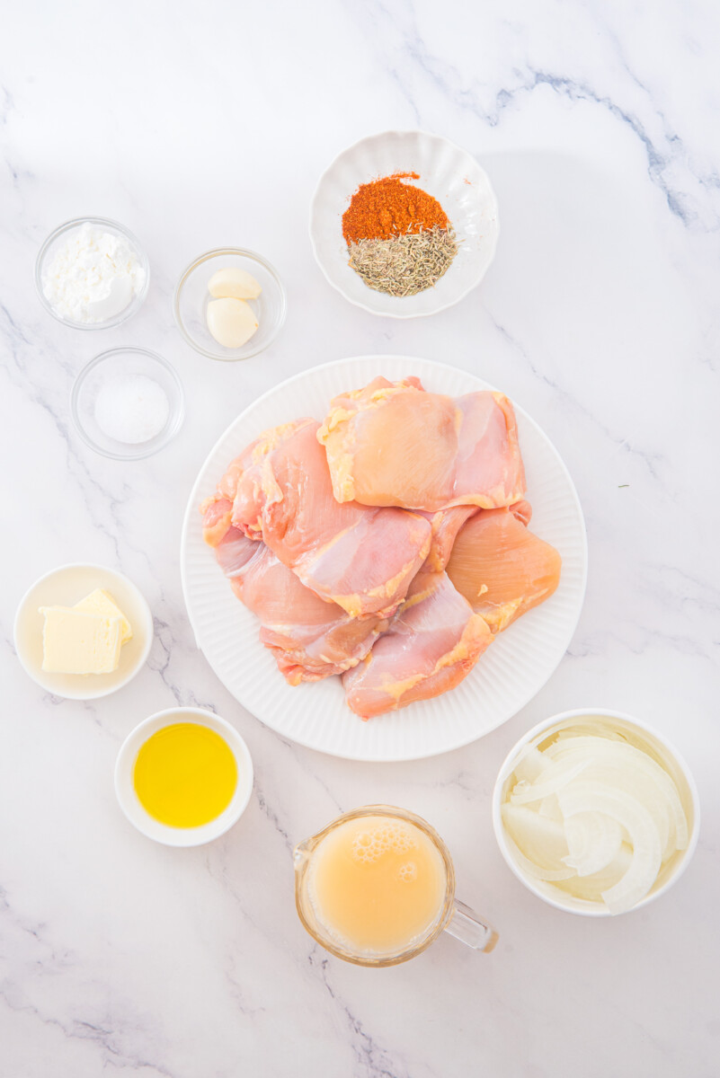 Smothered chicken ingredients laid out - raw chicken breasts with paprika, oats, butter, cream, and broth on marble counter