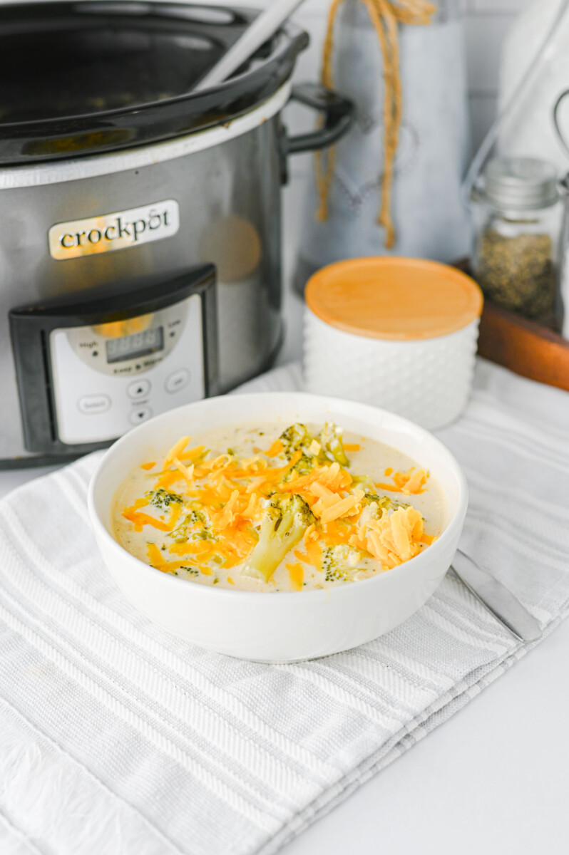 Crock Pot broccoli cheddar soup served in white bowl with cheddar cheese and broccoli garnish, slow cooker in background
