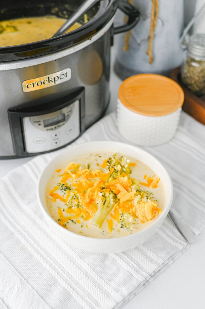 Crockpot broccoli cheddar soup in white bowl with cheese and broccoli garnish, slow cooker in background