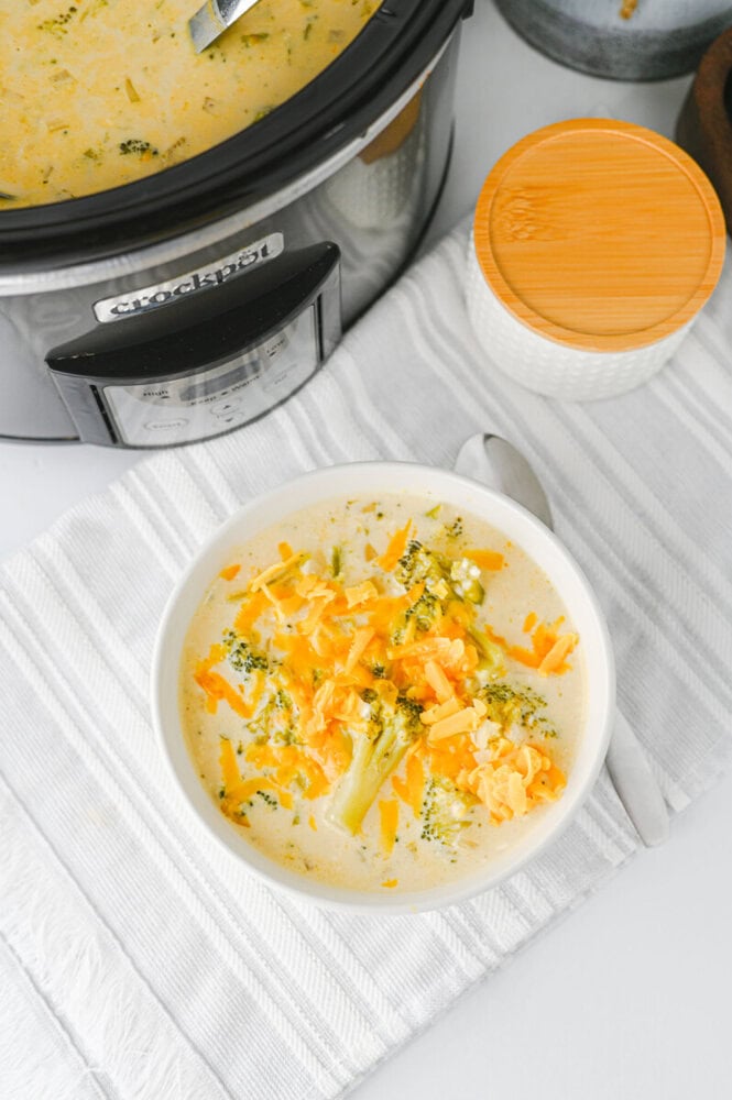 Crock-Pot broccoli cheddar soup served in a white bowl with cheddar cheese and herb garnish, slow cooker in background