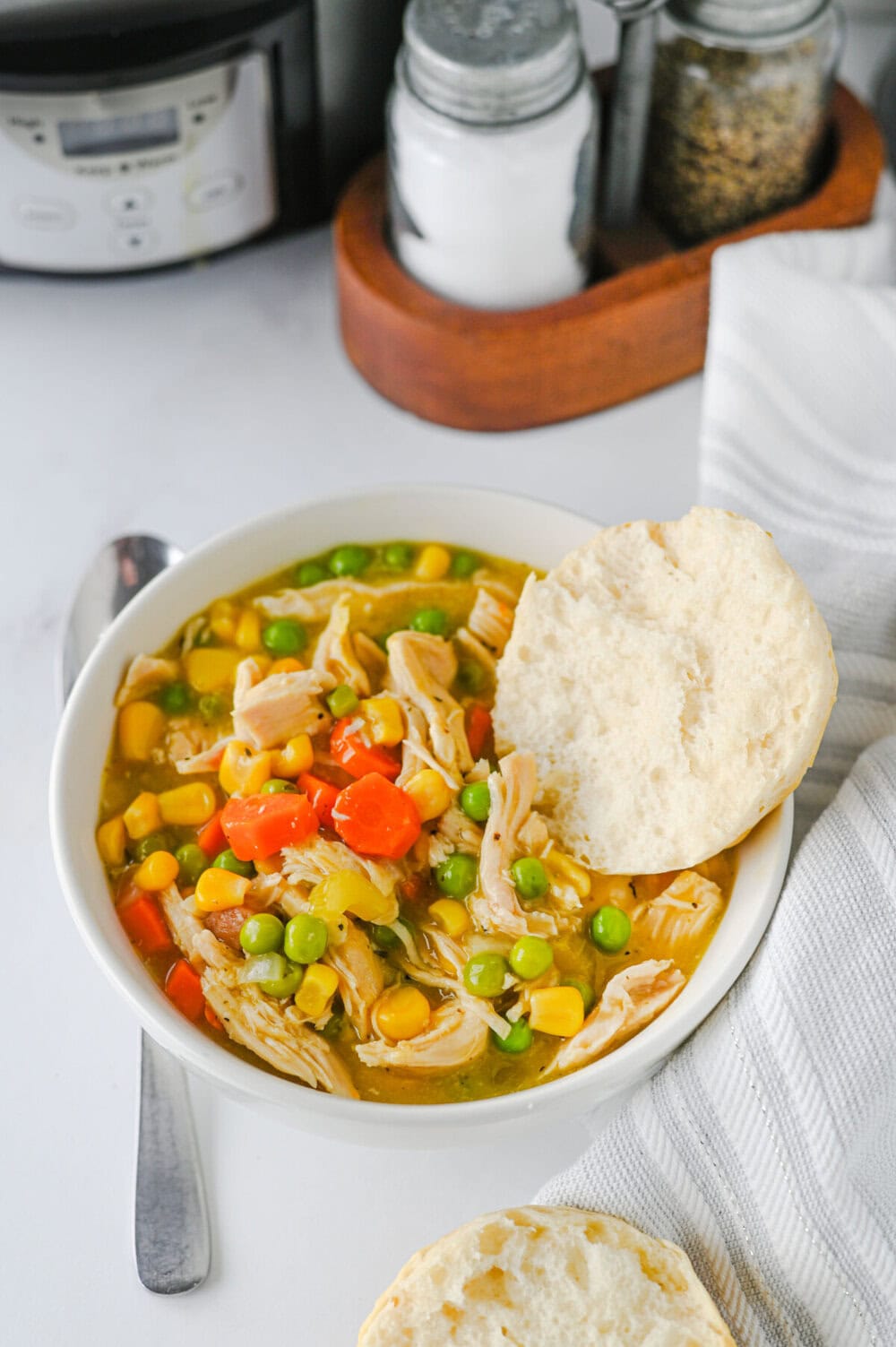 Crock Pot chicken and biscuits stew in white bowl with golden biscuit and vegetables including corn, peas, and red peppers