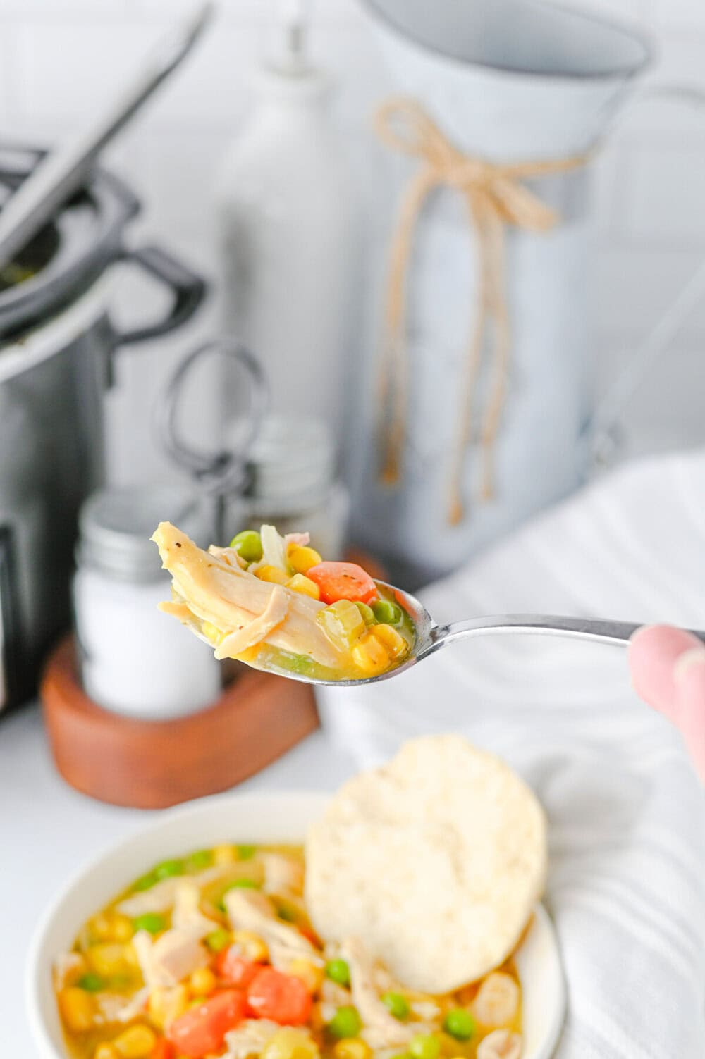 Crock pot chicken and biscuits served in a white bowl with a spoonful held up showing chunks of chicken, vegetables, and creamy sauce