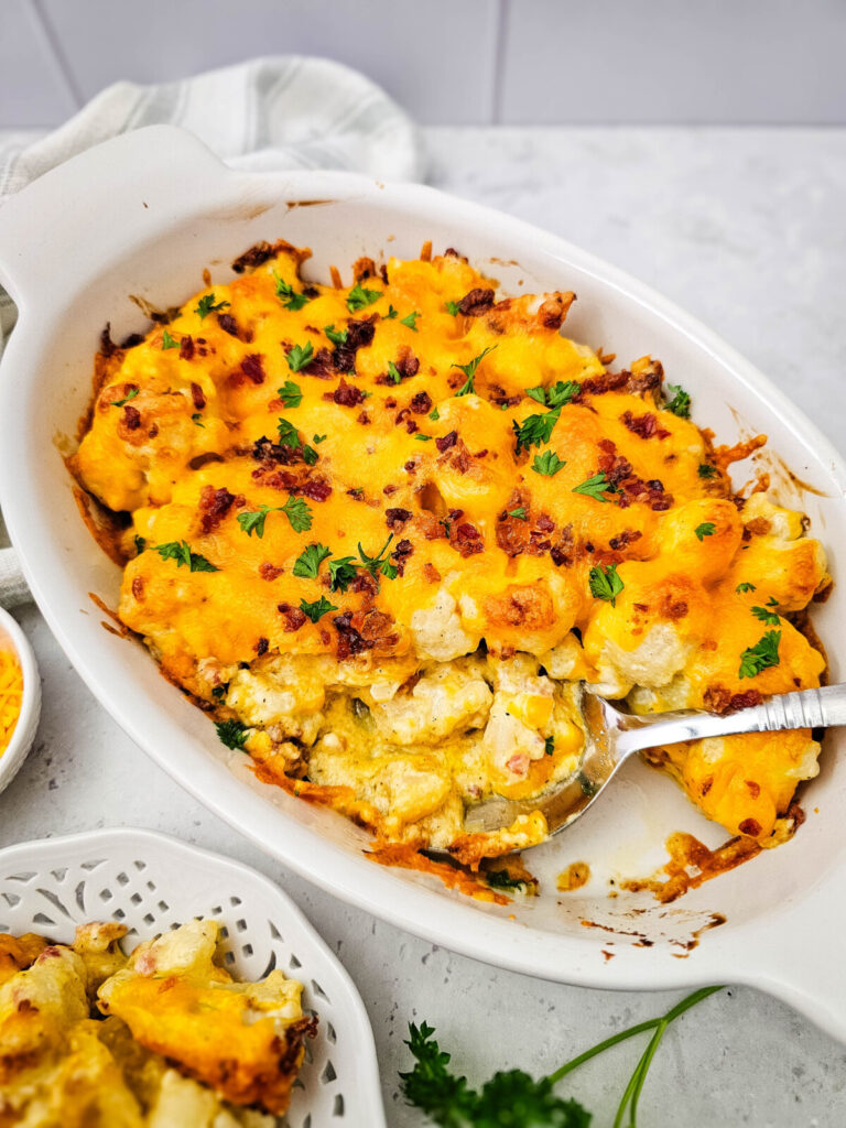 Baked cauliflower casserole with cheddar cheese and bacon bits topped with fresh parsley in white dish