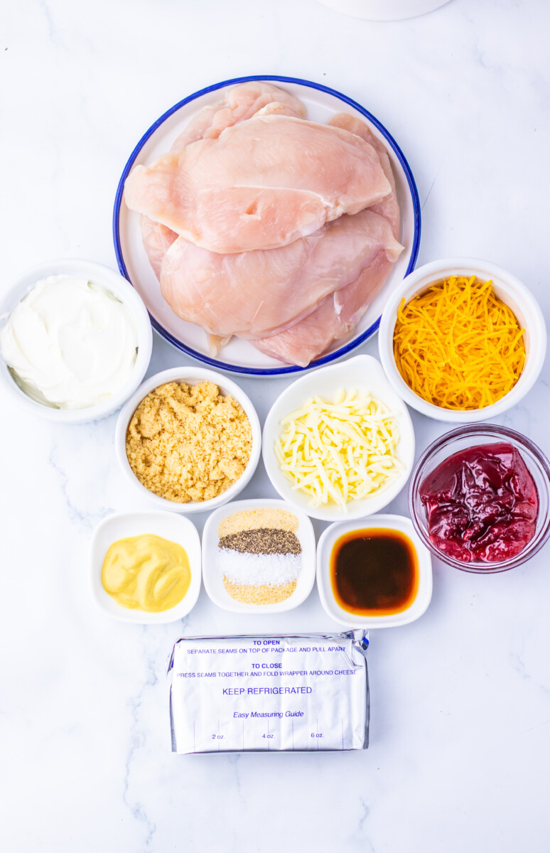 Ingredients for cranberry cream cheese chicken bake laid out in bowls including raw chicken breasts, cheeses, breadcrumbs, and sauce