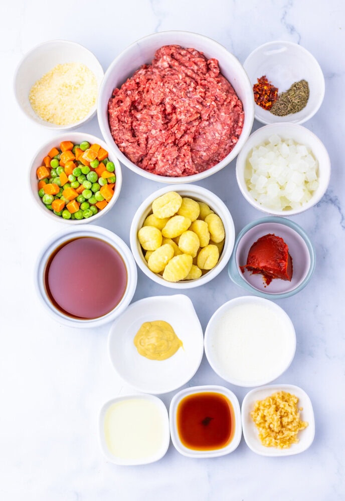 Shepherd's pie ingredients laid out in bowls including ground beef, gnocchi, peas and carrots, onions, seasonings, and sauces