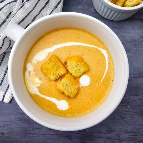 Creamy pumpkin soup in white bowl topped with croutons and cream swirl, served on dark surface with striped napkin