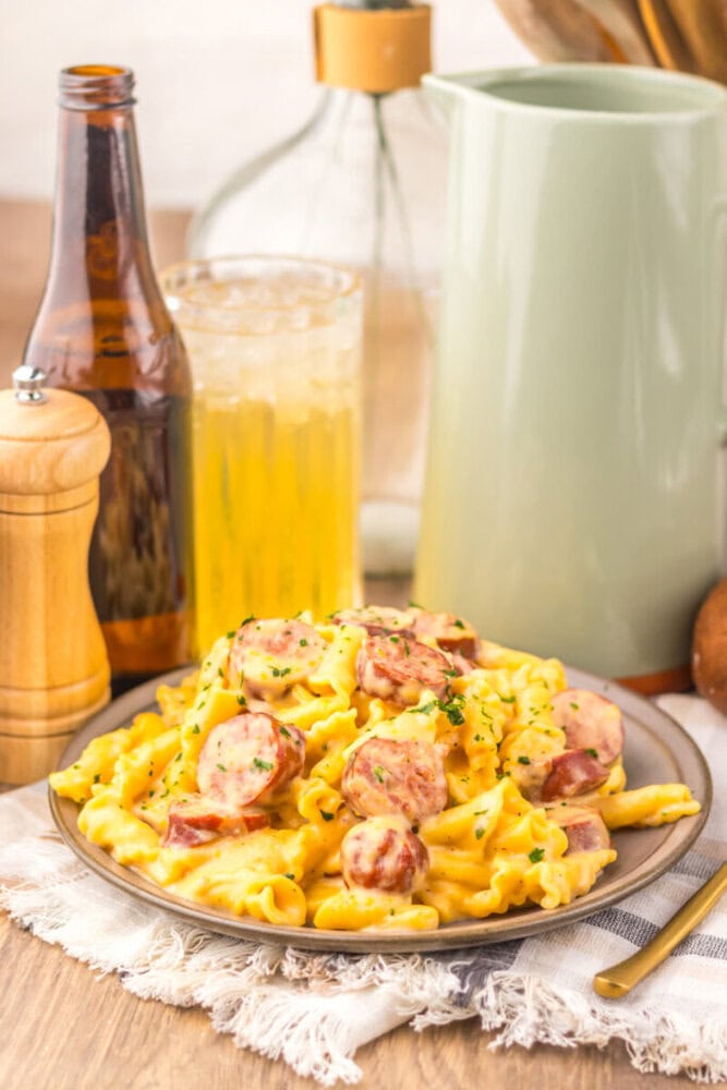 Slow cooker beer cheese sausage pasta with sliced sausage on gray plate, beer bottle and cream pitcher in background