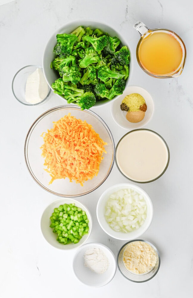 Crock pot broccoli cheddar soup ingredients prepped and arranged in bowls including broccoli florets, shredded cheddar cheese, diced onions and celery, broth, and cream