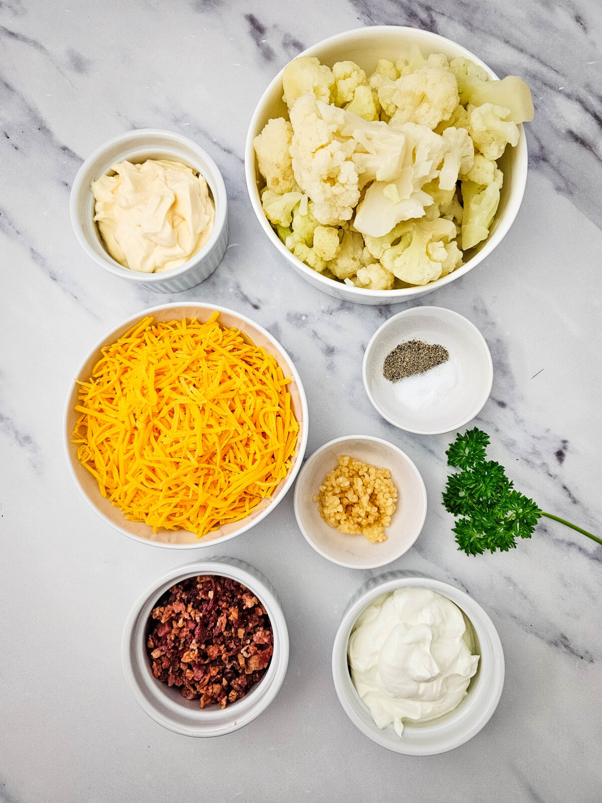 Cauliflower casserole ingredients laid out in bowls including cauliflower, cheddar cheese, mayo, sour cream, garlic, seasonings, bacon and parsley