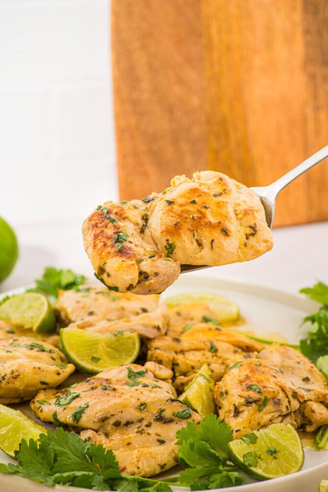 Cilantro lime chicken on a white plate garnished with fresh cilantro and lime, with a fork holding a piece overhead