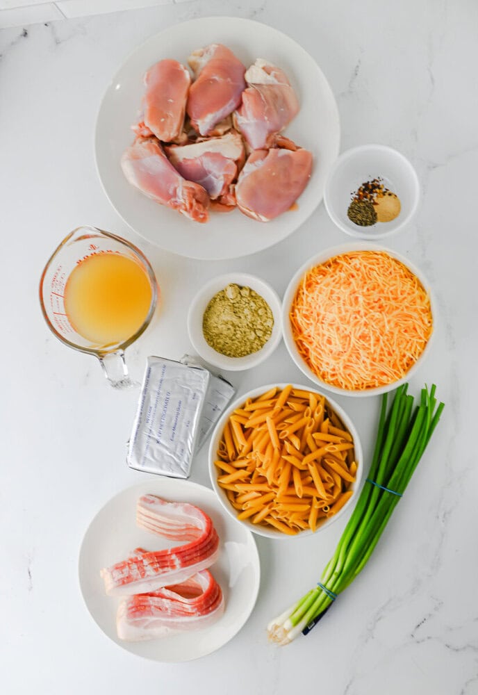 Crockpot Crack Chicken Pasta ingredients arranged overhead including raw chicken, bacon, cheese, pasta, and seasonings on white marble