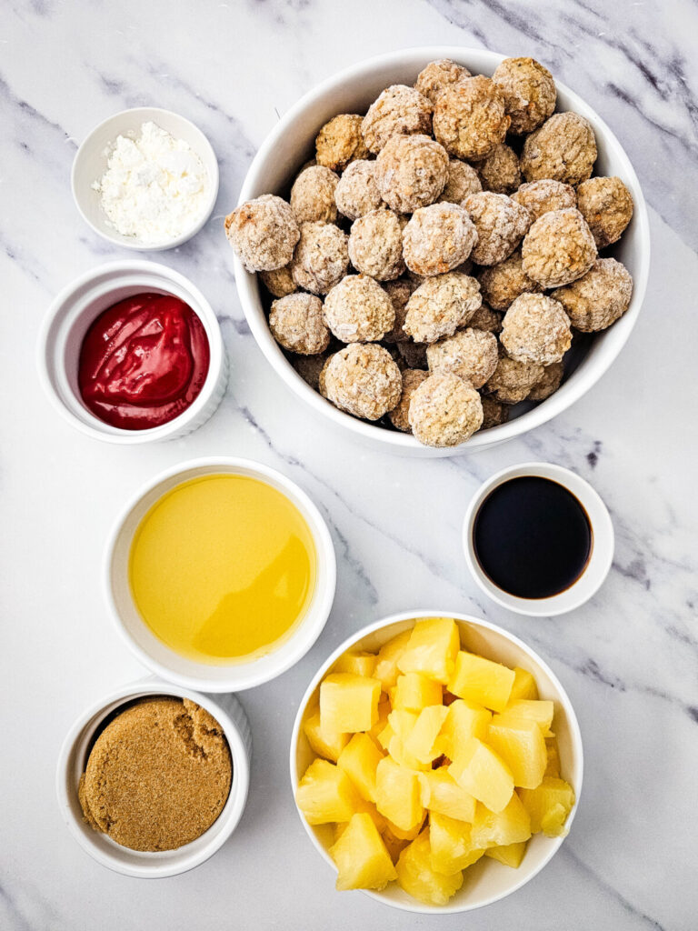 Crockpot sweet and sour meatballs ingredients laid out in bowls including meatballs, pineapple, sauces, and spices on marble
