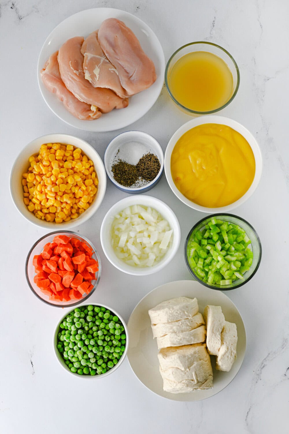 Crock pot chicken and biscuits ingredients laid out in bowls including chicken breasts, frozen vegetables, broth, and biscuit dough
